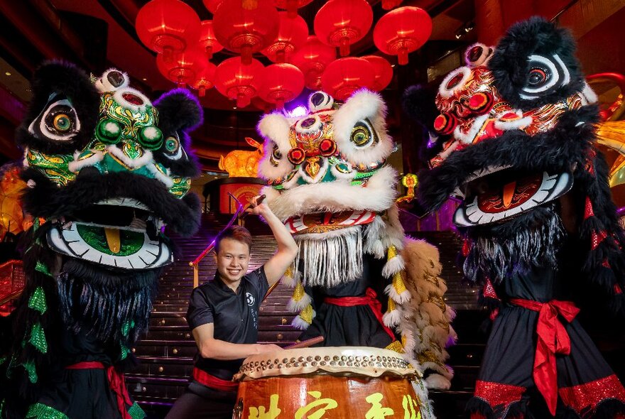 A smiling performer standing in front of a large drum with three ornately decorated Chinese dragon and lion puppets behind him.