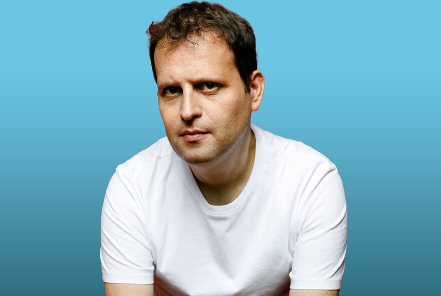 Writer Adam Kaye, in a white t-shirt against a blue background, sitting forward looking serious with a heavy brow.