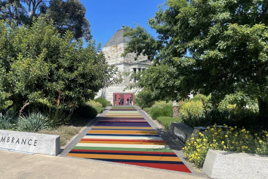 Striped colours, representing Australian military medal ribbons, along a pathway leading up to the Shrine of Remembrance, with leafy green trees alongside.