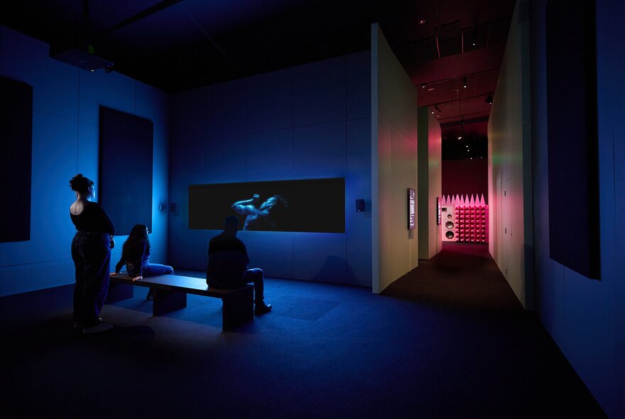 Installation in a dimly lit room with deep blue lighting at ACMI, with a few people silhouetted as they view a horizontal, rectangular screen. 