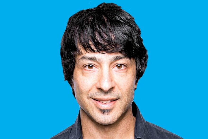 Comedian Arj Barker head shot against a blue background.