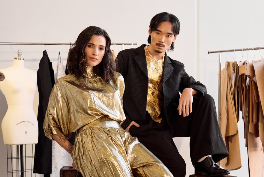Two fashion models striking a pose inside a garment studio, racks of hanging patterns and a mannequin in the background.