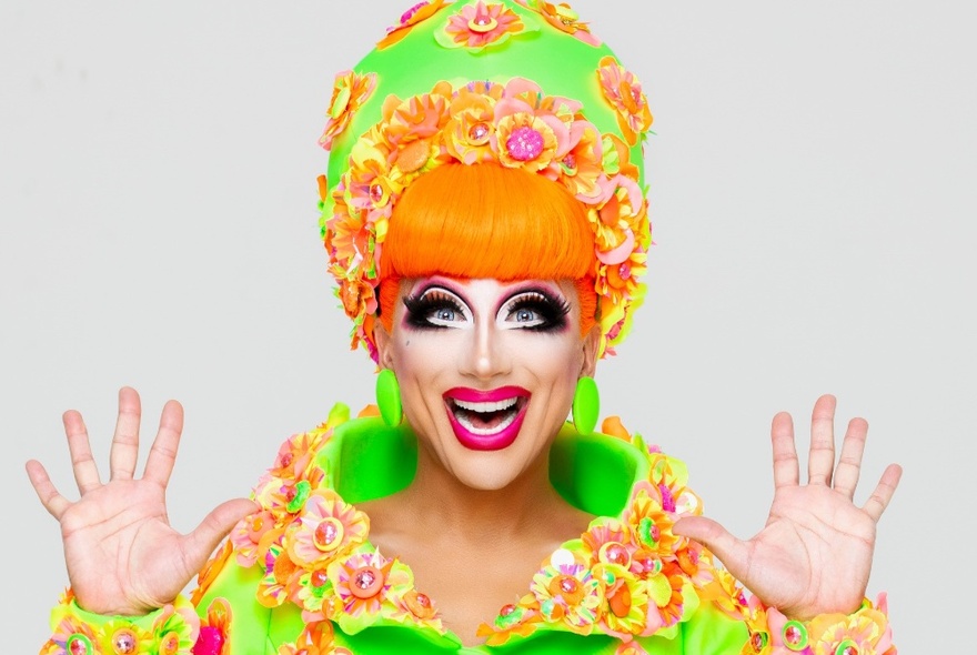 Drag queen Bianca Del Rio in full makeup and an orange and lime green outfit with flowers, her hands up.