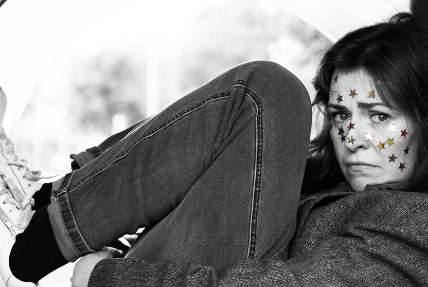 Black and white image of adult with legs folded up near her chest, her face covered in coloured star stickers and with a puzzled expression on her face.