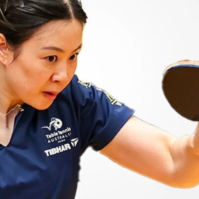 Woman concentrating while playing table tennis, she holds a bat in one hand and is wearing a navy blue t-shirt with logos on it.