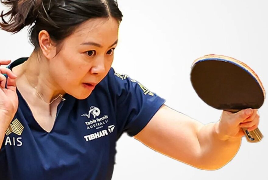 Woman concentrating while playing table tennis, she holds a bat in one hand and is wearing a navy blue t-shirt with logos on it.
