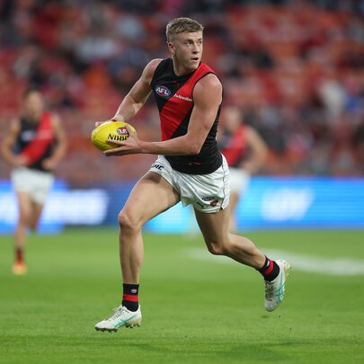 An Essendon AFL football player running while holding the ball during a match, the blurred stadium crowd in the background.