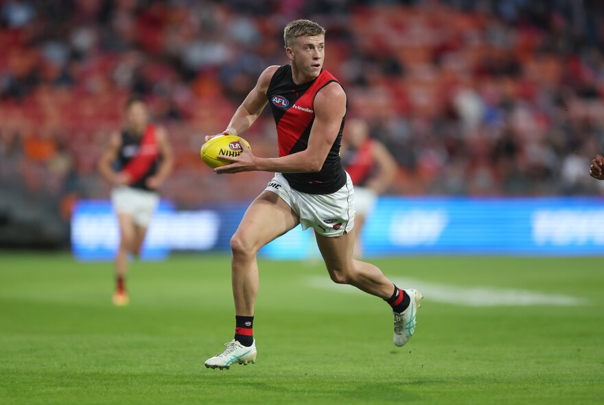 An Essendon AFL football player running while holding the ball during a match, the blurred stadium crowd in the background.