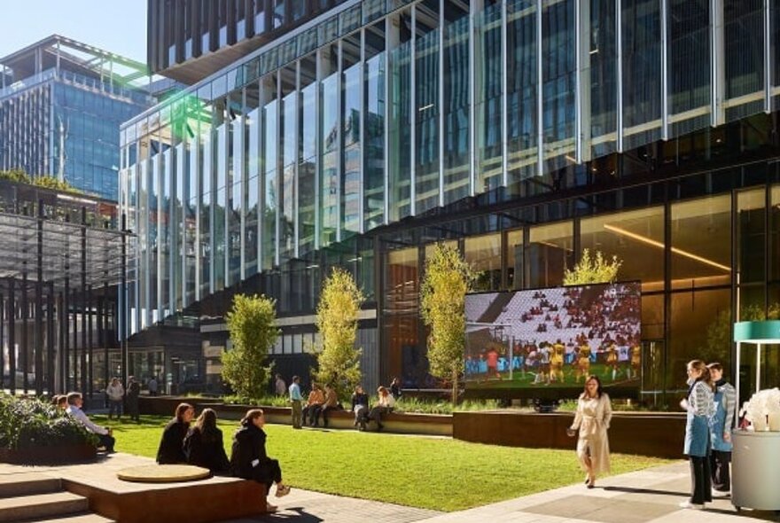 The sunny forecourt of a glass covered office building, with people standing around, walking or sitting.