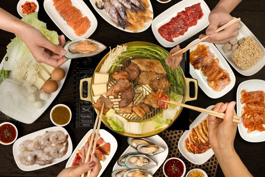 Hands reaching for food from an array of Asian dishes with chopsticks.