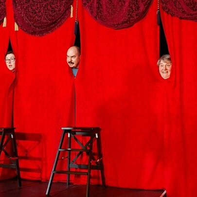 Three faces poking through gaps in a long red curtain on a stage with stools in front of the stage.