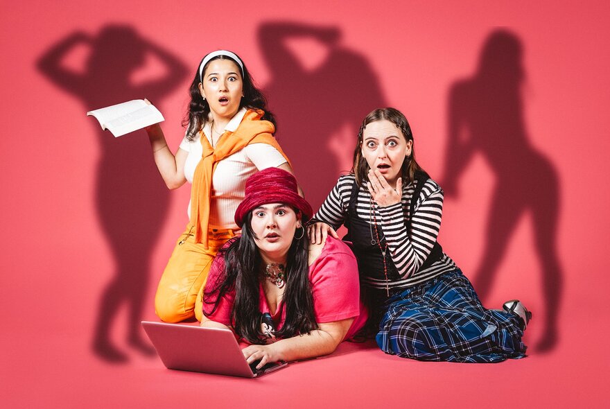Three young women in colourful clothes sitting, lying or crouching on the floor, one with a laptop in front of her; sexually suggestive shadows on the wall behind them.