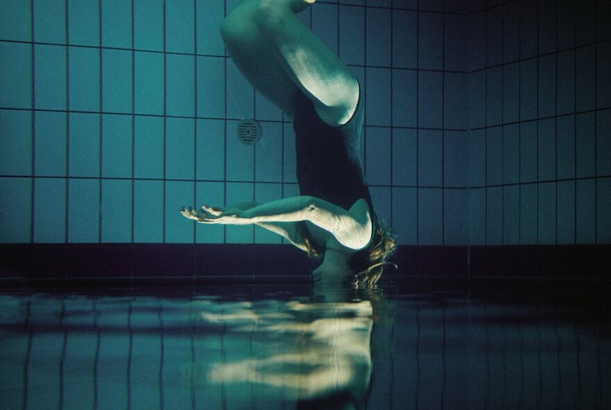 An upside down shot of a woman underwater wearing a black bathing suit, listening to underwater music.