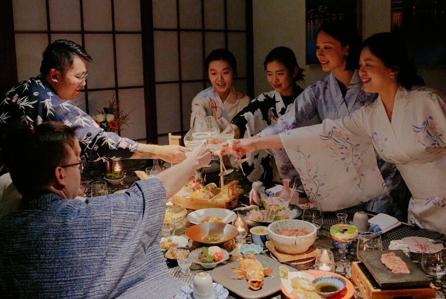 People dressed in kimono clinking glasses over  a table filled with dishes in a private Japanese restaurant space.