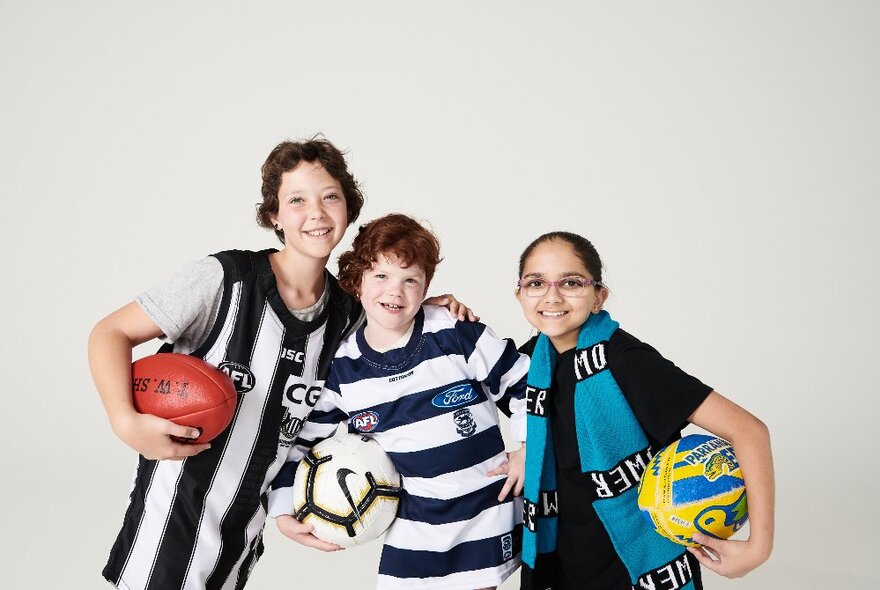 Three kids wearing footy colours, one holding an AFL football, the other two holding soccer balls.