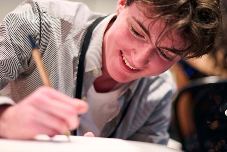 A smiling teenager holding a pencil against a piece of paper, concentrating on what he his doing.