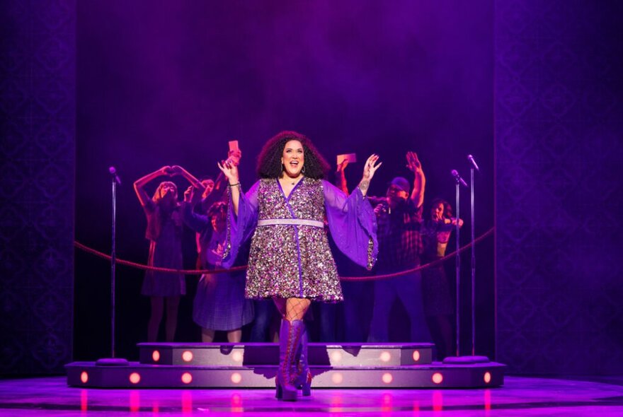 A woman on a purple-lit stage in a sparkly dress, singing.