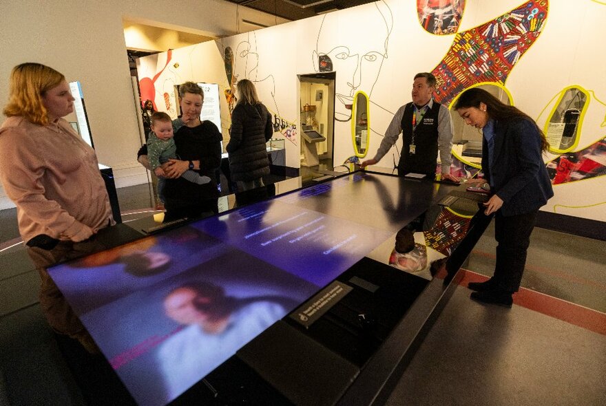 People on a tour at the Immigration Museum, looking over a table with a video display. 