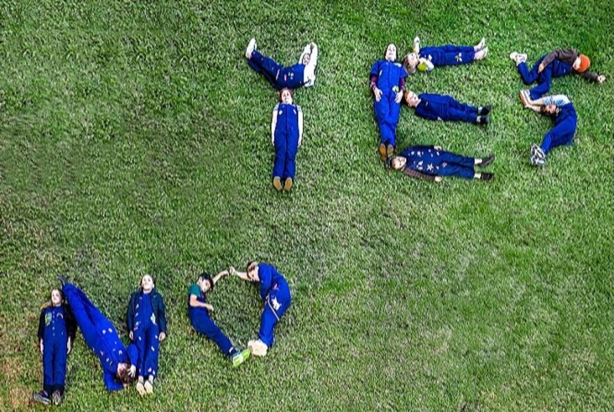 People lying on green grass forming the words Yes and No, wearing blue uniforms, viewed from above.