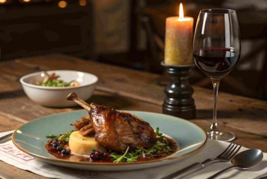 A gourmet duck dish on a duck-egg blue plate, with a candle and glass of wine on a rustic farm-style wooden table.