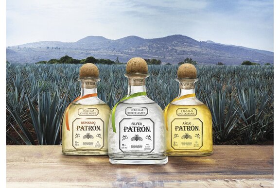 Three tequila bottles resting on a wooden table in front of an agave plantation, mountains in the distance.