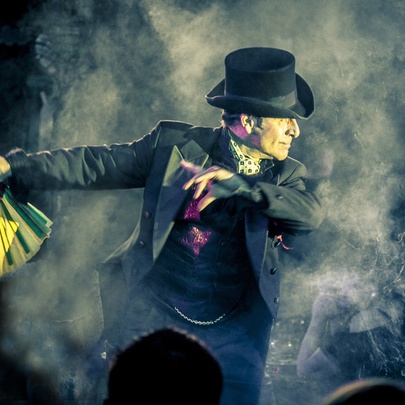 A male magician in a top hat and suit on a smoky stage, holding a yellow and green fan and performing a trick.