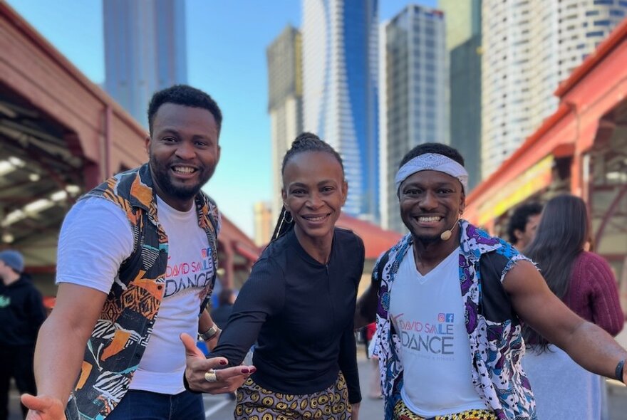 Three people from an African dance group, smiling and posing at the Queen Victoria Market with city buildings behind them. 