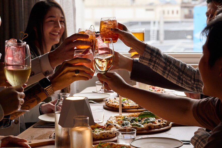 A group of patrons all holding drinks together in a cheers above a table laden with pizza.