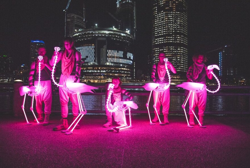 Performers holding illuminated pink lit-up flamingos on a rooftop with city buildings at night.