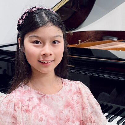 A smiling young teenager, with straight black hair and wearing a pastel coloured dress, osed in front of a grand piano.