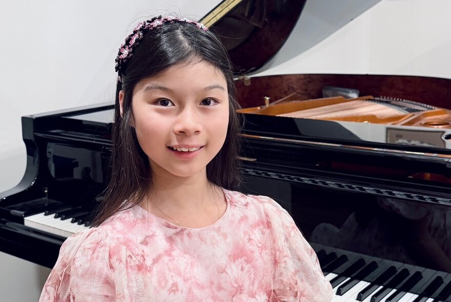 A smiling young teenager, with straight black hair and wearing a pastel coloured dress, osed in front of a grand piano.