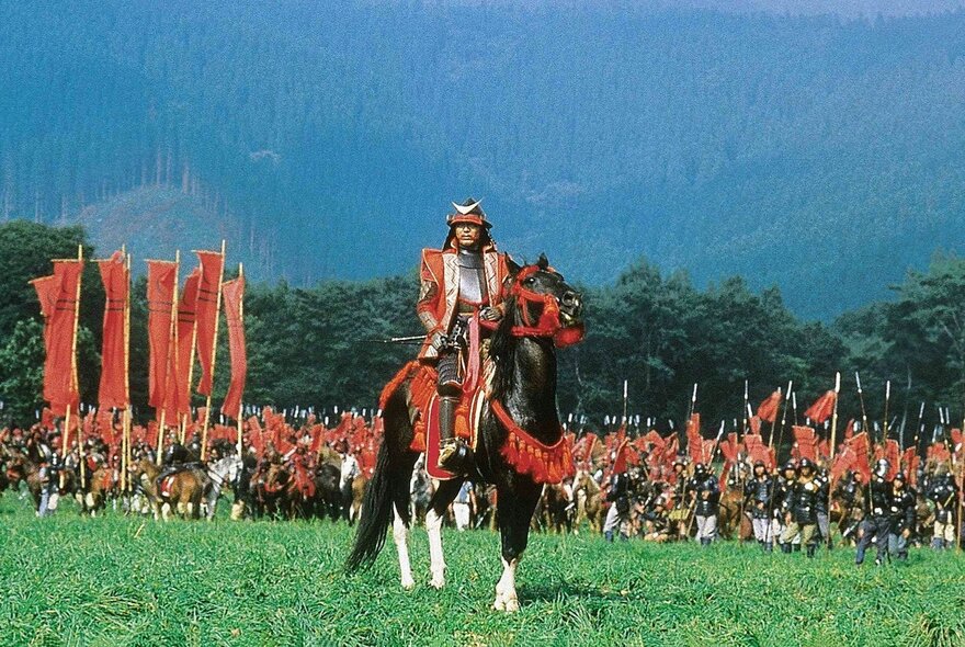A still from Akira Kurosawa's film Ran, with a leader on a horse with troops on horseback behind him, all with red flags and uniforms. 