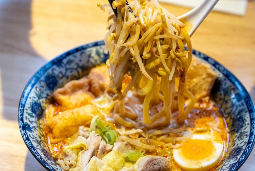 Cutlery lifting noodles from a bowl of Malaysian style soup, half a boiled egg on top. 