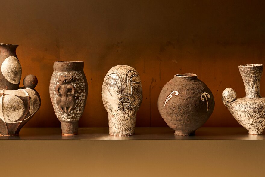 Five contemporary ceramics on display on a shelf in a gallery, against an ochre coloured wall.