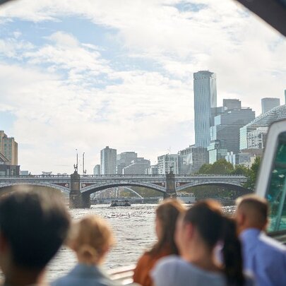 People standing on a boat looking over the river at a view of Princes Bridge and city buildings.