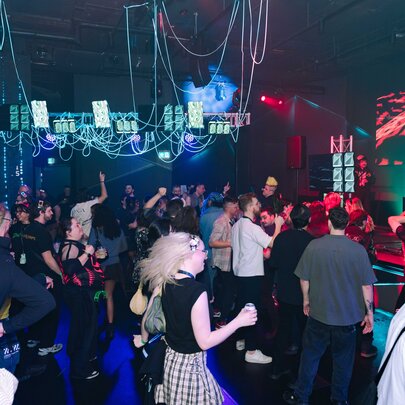 A crowded, dimly-lit venue filled with people dancing, and cables and small green screens hanging from the ceiling.