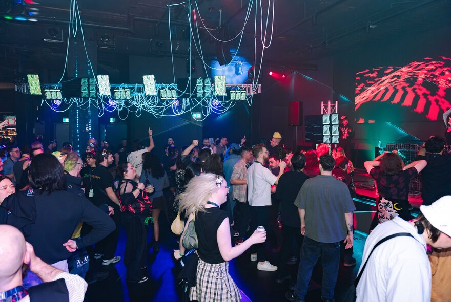 A crowded, dimly-lit venue filled with people dancing, and cables and small green screens hanging from the ceiling.