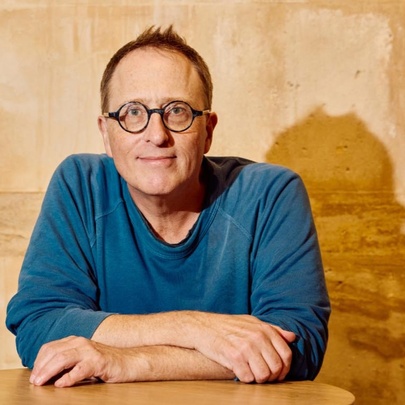 The author and journalist Jon Ronson, a middle-aged man wearing round glasses and a blue top, looking directly at the viewer with a neutral expression, forearms resting on a table, posed against a light tan-coloured wall.
