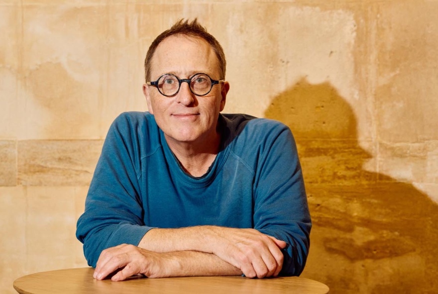 The author and journalist Jon Ronson, a middle-aged man wearing round glasses and a blue top, looking directly at the viewer with a neutral expression, forearms resting on a table, posed against a light tan-coloured wall.