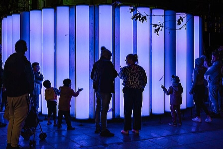 Blue-lit pipes in the forecourt of the Melbourne Arts Centre, with people standing around and touching them.