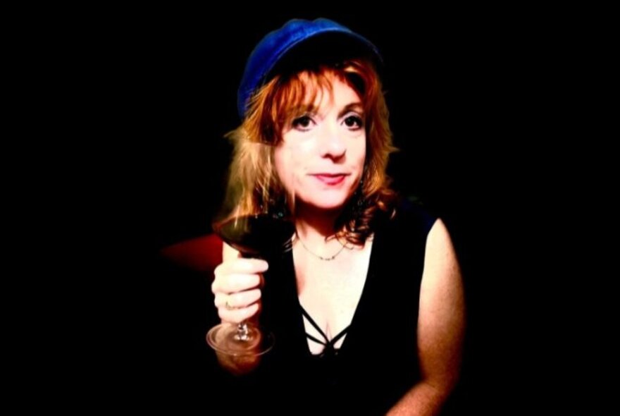 Singer and pianist Katie Pryce wearing a blue cap and a black vest, holding a glass of red wine, posed against a black background.