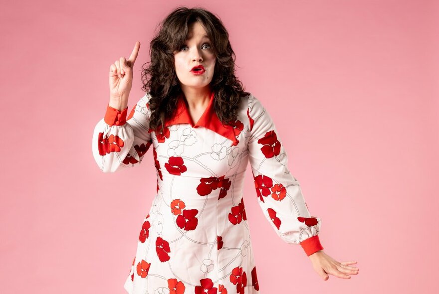 Singer and performer Alice Tovey in a white dress with a large red floral pattern on it, standing in front of a pink backdrop.