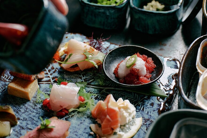 Finely styled sushi items on a patterned ceramic plate with dark bowls. 