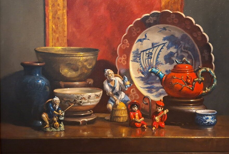 Still life oil painting of ceramic bowls, a platter, porcelain figurines and a bronze vessel arranged on a wooden sideboard.