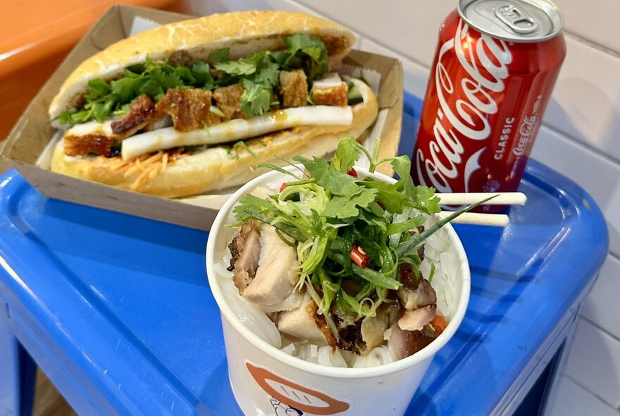 Banh mi sandwich, cup of Asian soup and can of Coca-Cola on a blue stool.