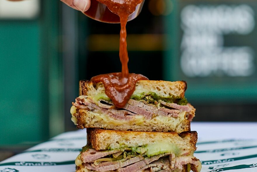 A red hot sauce being drizzled over the top of a stacked cut-beef sandwich.