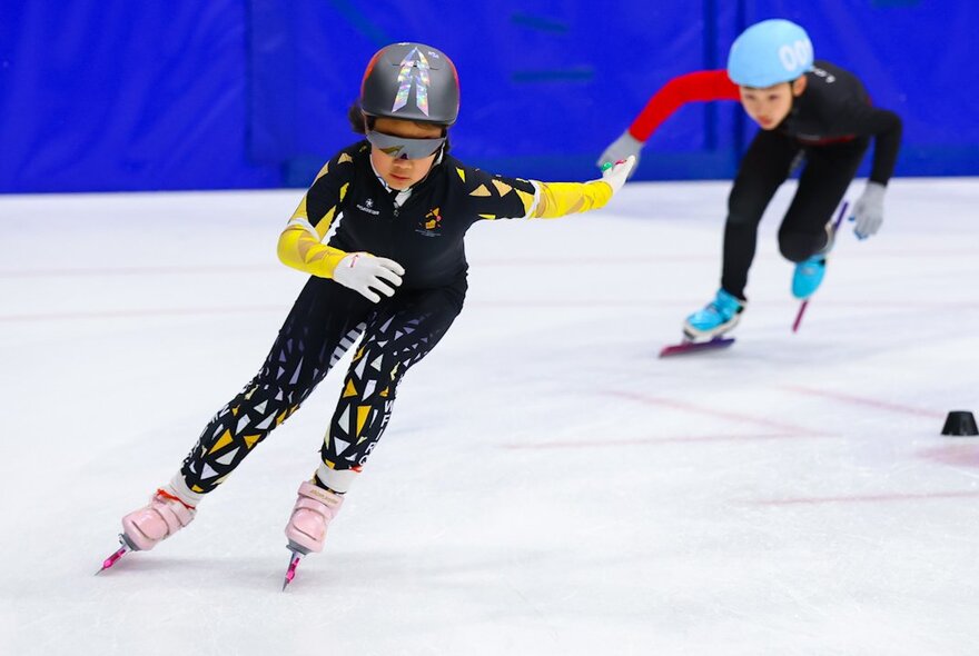Two young athletes short-track speed skating on an indoor ice rink.