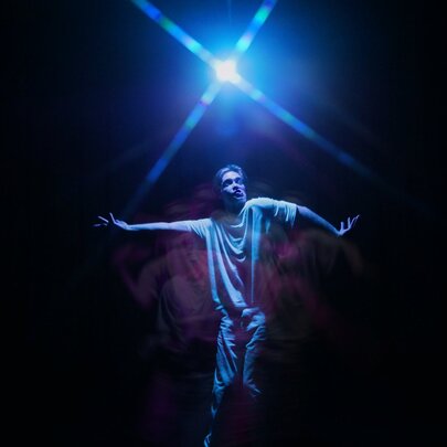 Physical theatre performer Daniel Nodder with his arms outstretched and holding a pose, on a dark stage under a single white light globe.