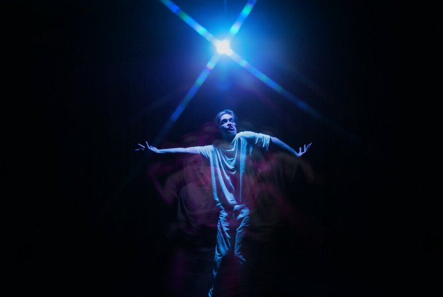 Physical theatre performer Daniel Nodder with his arms outstretched and holding a pose, on a dark stage under a single white light globe.