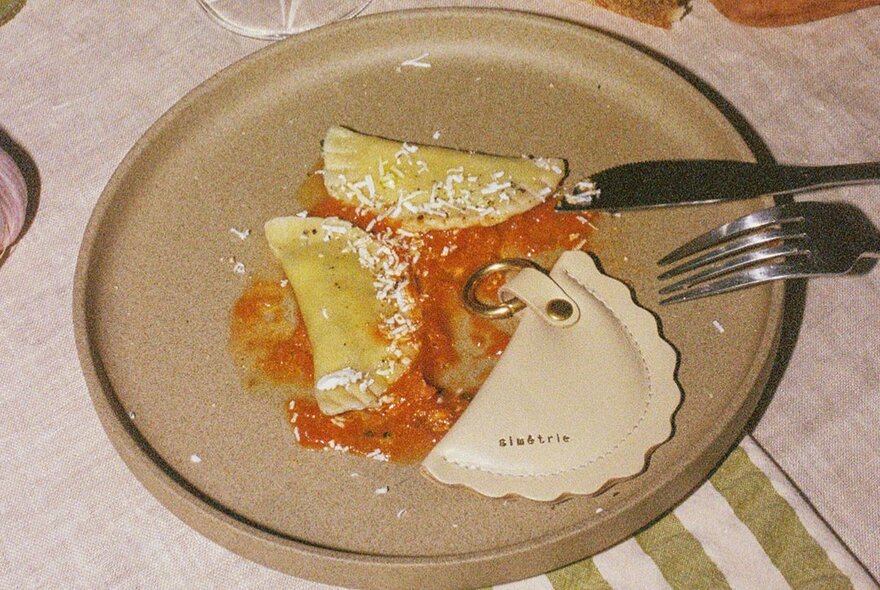 A plate with two pasta agnolotti in a tomato sauce, with a small leather coin purse in the same half moon shape as the agnolotti also on the plate.